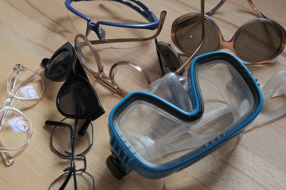 Auf einer hölzernen Tischplatte sind mehrere Brillenmodelle angeordnet. Darunter auch eine Sonnen- und eine Taucherbrille.
